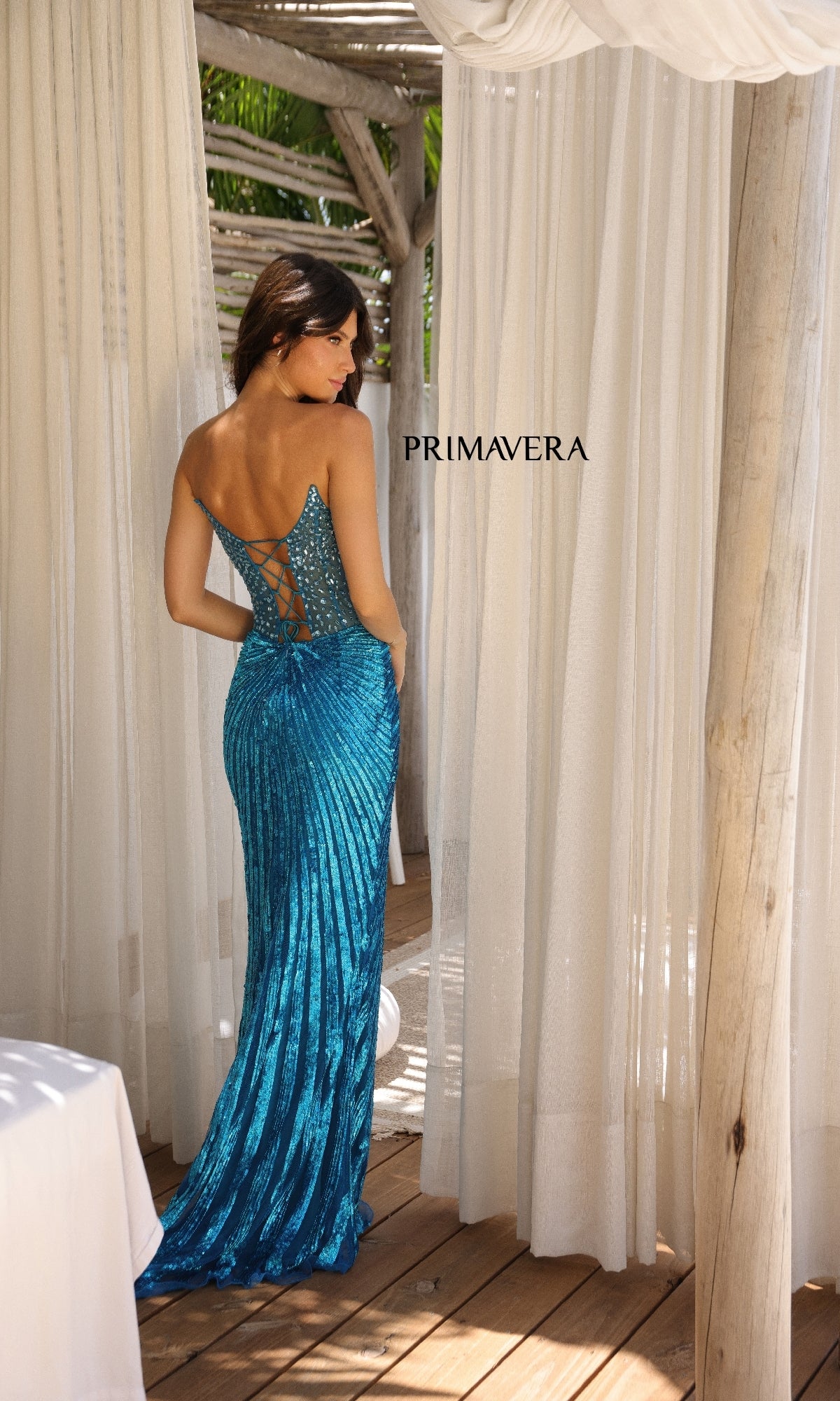 A woman wearing the Strapless Long Sequin Prom Dress, Primavera 4304, in sparkling blue with a lace-up back stands by white curtains under a wooden pergola. The word